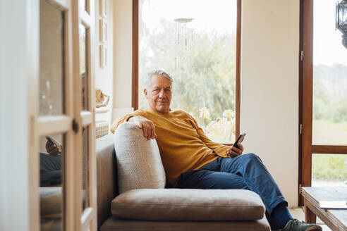 Smiling senior man sitting with mobile phone on sofa at home - JOSEF16419