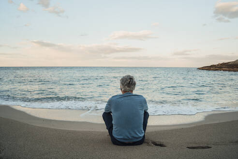 Senior man sitting on sand in front of sea at beach - JOSEF16482