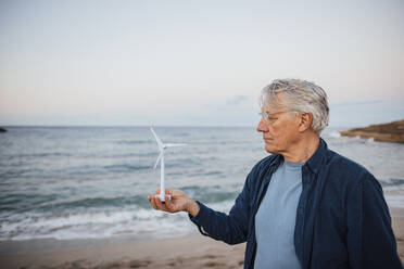 Senior man standing with wind turbine model in front of sea - JOSEF16500