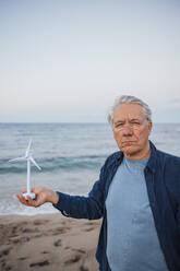 Man standing with wind turbine model in front of sea at beach - JOSEF16501