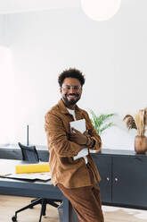 Happy businessman with laptop leaning on desk in office - PNAF04885