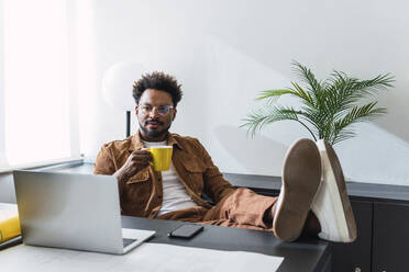 Geschäftsmann mit Kaffeetasse am Schreibtisch sitzend vor einer Wand - PNAF04890