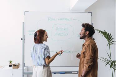 Lächelnde Geschäftsfrau im Gespräch mit einem Kollegen, der vor einem Whiteboard steht - PNAF04929