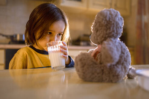 Junge schaut Teddybär auf Tisch an und trinkt Milch - ANAF00962