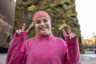 Smiling mature woman in pink sweater showing peace sign - JCCMF09068