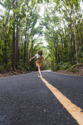 Young woman walking on road marking in front of trees - PNAF04958