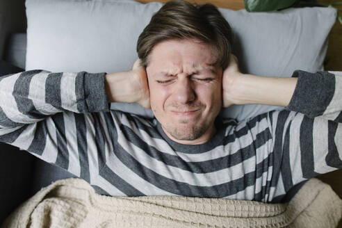 Frustrated man covering ears lying on bed at home - TYF00695