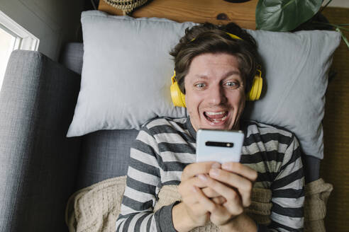 Aufgeregter Mann mit Smartphone und Kopfhörern, der zu Hause auf dem Bett liegt - TYF00697