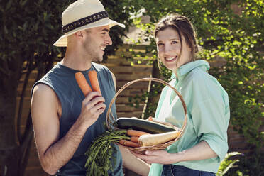 Glückliches junges Paar steht mit einem Korb voller Gemüse im Hinterhof - PWF00678
