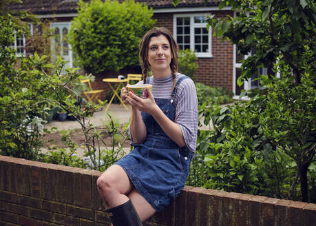 Thoughtful woman with mug sitting on fence in back yard - PWF00711