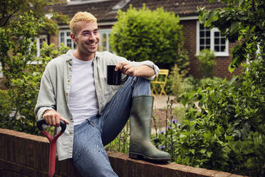 Happy young man wearing rubber boot sitting on fence in back yard - PWF00714