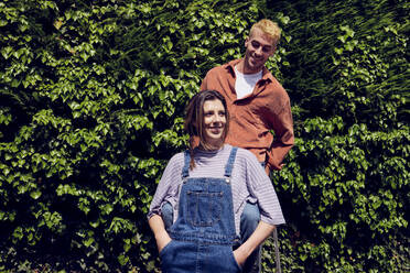 Smiling woman with hands in pockets standing by boyfriend near hedge - PWF00720