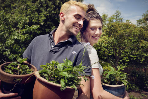 Happy couple standing back to back holding potted plants - PWF00737