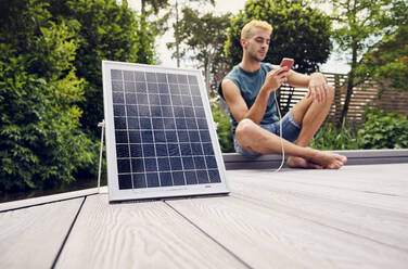 Junger Mann sitzt auf der Terrasse und benutzt sein Smartphone, das von einem Solarpanel aufgeladen wird - PWF00755
