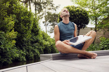 Junger Mann, der ein Solarpanel hält und im Schneidersitz auf der Terrasse sitzt - PWF00756