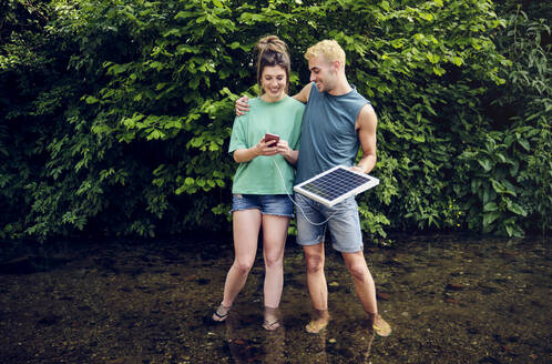 Glückliches Paar, das ein Smartphone benutzt, das von einem Solarpanel aufgeladen wird, das im Fluss steht - PWF00758