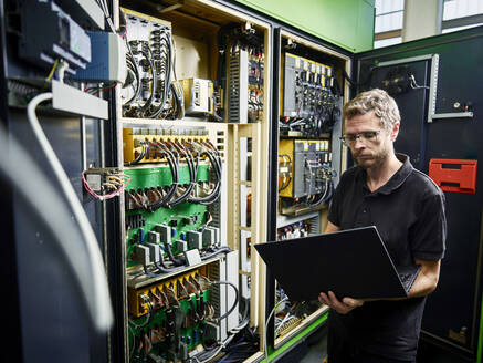 Technician using laptop at machine in modern factory - CVF02285