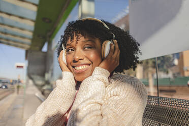 Happy woman wearing wireless headphones sitting at station - JCCMF09077