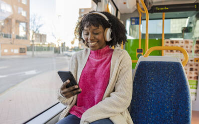 Happy woman wearing wireless headphones using smart phone in tram - JCCMF09079