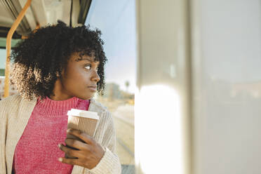 Junge Frau mit Kaffeetasse schaut durch ein Fenster in der Straßenbahn - JCCMF09085