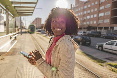 Glückliche junge Frau mit Smartphone am Bahnhof stehend - JCCMF09093