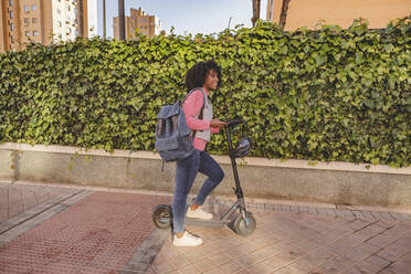 Junge Frau mit Elektro-Scooter auf dem Gehweg stehend - JCCMF09095