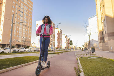 Glückliche junge Frau, die auf einem elektrischen Roller auf dem Gehweg fährt - JCCMF09105