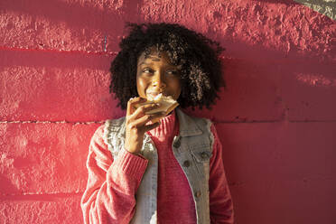 Smiling young woman with sandwich standing in front of red wall - JCCMF09129