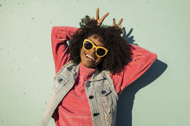 Cheerful young woman wearing sunglasses showing peace gesture in front of wall - JCCMF09138