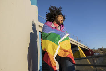 Fröhliche junge Frau, eingewickelt in eine Regenbogenfahne, genießt einen sonnigen Tag - JCCMF09143