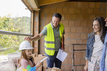 Ein lächelnder Bauunternehmer setzt einem Mädchen mit Familie auf der Baustelle einen Schutzhelm auf den Kopf - EIF04203