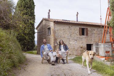 Happy mature couple sitting on chair with dog in front of house - EIF04233