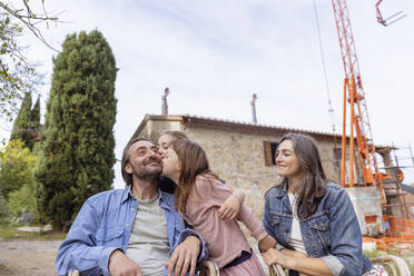 Girls kissing father with mother sitting in front of house - EIF04236