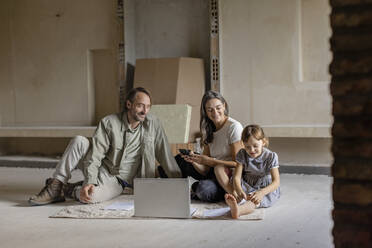 Happy family sitting with laptop and documents on rug at home - EIF04324