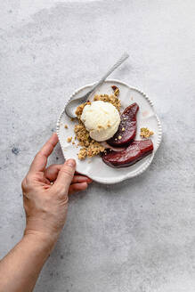 Woman's hand holding plate of pears in red wine with ice cream and granola - FLMF00924