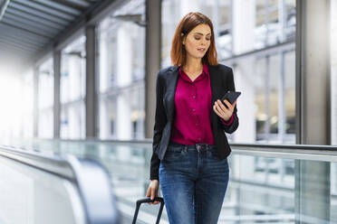 Businesswoman using smart phone pulling luggage at airport - DIGF19706