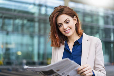 Smiling redheaded businesswoman reading newspaper - DIGF19746