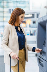 Lächelnde Geschäftsfrau mit Gepäck, die eine Kreditkarte am Geldautomaten benutzt - DIGF19750
