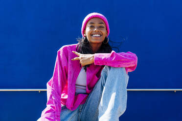 Glückliche junge Frau, die vor einer blauen Wand sitzend ein Friedenszeichen macht - OIPF03063
