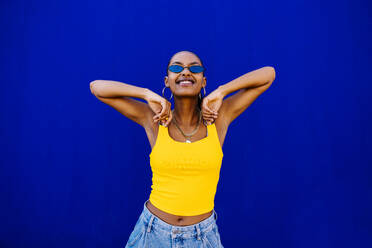 Glückliche junge Frau mit Sonnenbrille vor einer blauen Wand - OIPF03071