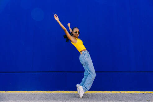 Young woman on tiptoes with arms raised by blue wall - OIPF03076