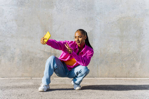 Smiling young dancer in squatting position taking selfie through mobile phone in front of wall - OIPF03115