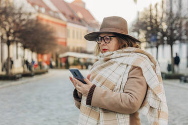 Senior woman using smart phone standing on street - VSNF00383