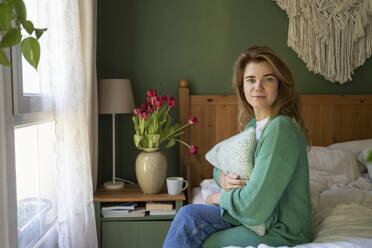 Smiling woman with pillow sitting on bed at home - SVKF01199