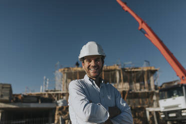 Happy architect standing with arms crossed at construction site - DMGF00977
