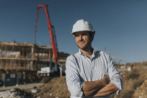 Nachdenklicher reifer Arbeiter mit verschränkten Armen auf einer Baustelle - DMGF00978