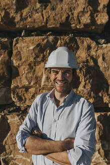 Glücklicher reifer Architekt mit Schutzhelm vor einer Backsteinmauer stehend - DMGF00981