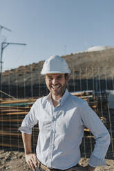 Happy mature architect wearing hardhat standing at construction site - DMGF00988