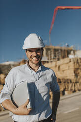 Happy architect standing with laptop at construction site - DMGF01001