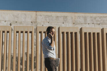 Businessman talking over smart phone standing by wall on sunny day - DMGF01003